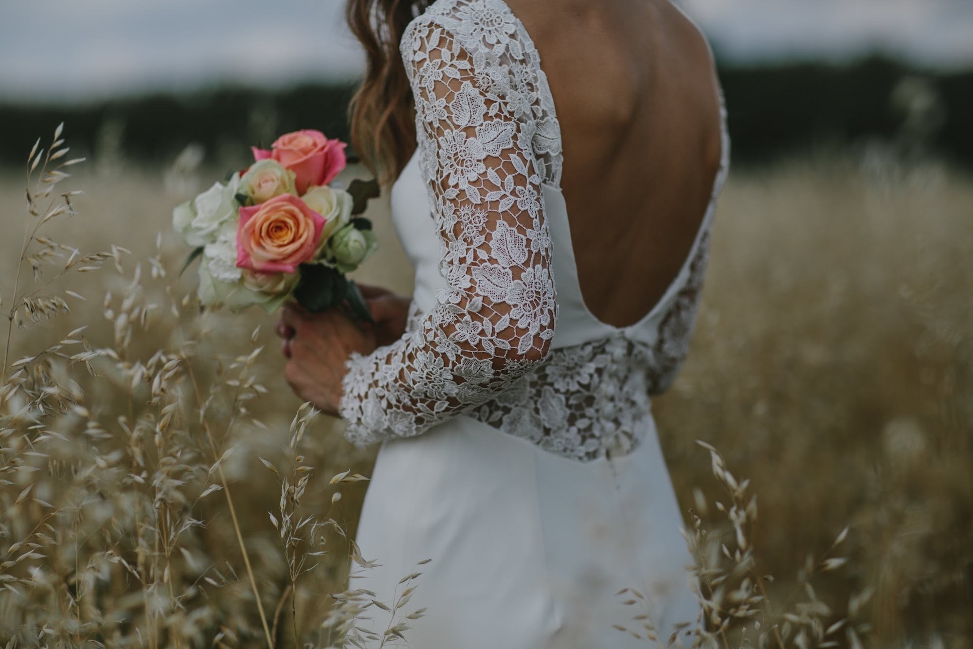Bride in a lace-backed dress holding a bouquet in a field of oat grass, Lesley Cutler Bridal Wear