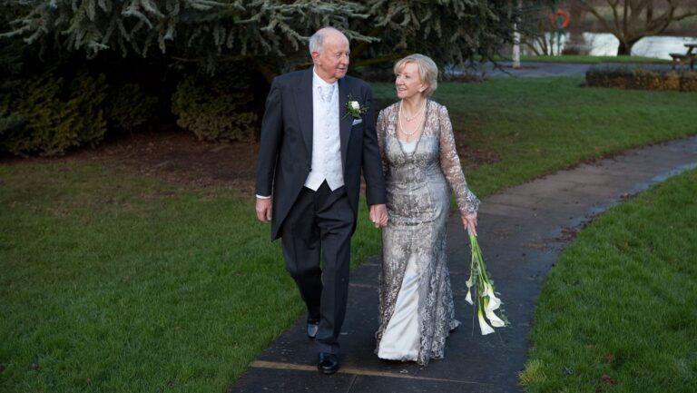 Couple walking on a garden path, the bride in a fitted lace gown, Lesley Cutler Bridal Wear