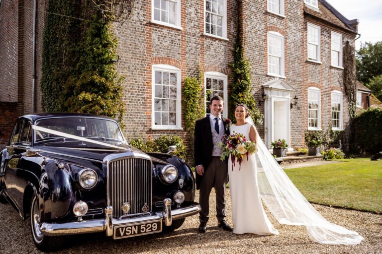 Bride and groom beside a vintage car in front of a brick house, Lesley Cutler Bridal Wear