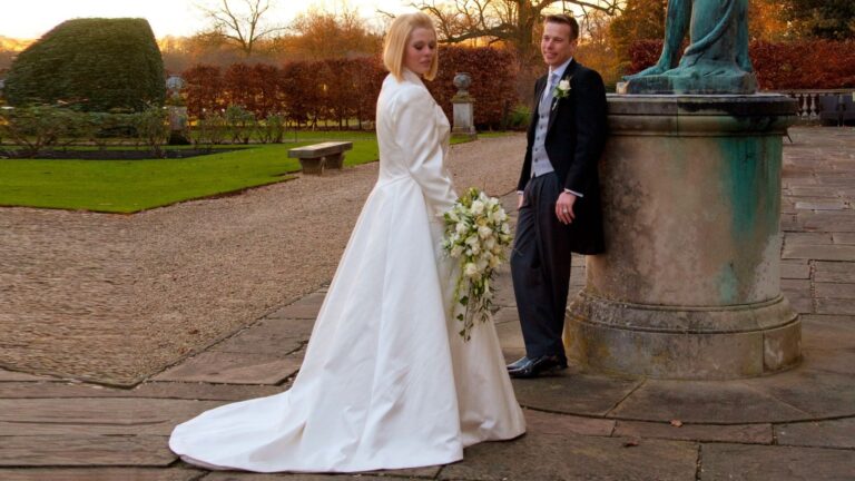 Bride in a long-trained gown beside a statue in a garden, Lesley Cutler Bridal Wear