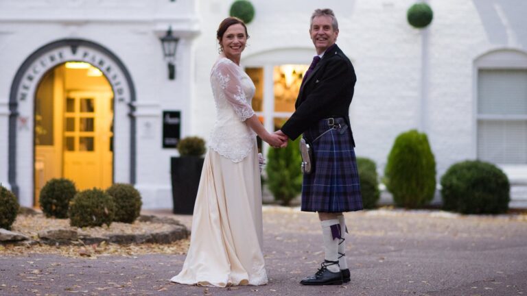 Couple holding hands outside a hotel, the bride in an ivory gown with lace sleeves, Lesley Cutler Bridal Wear