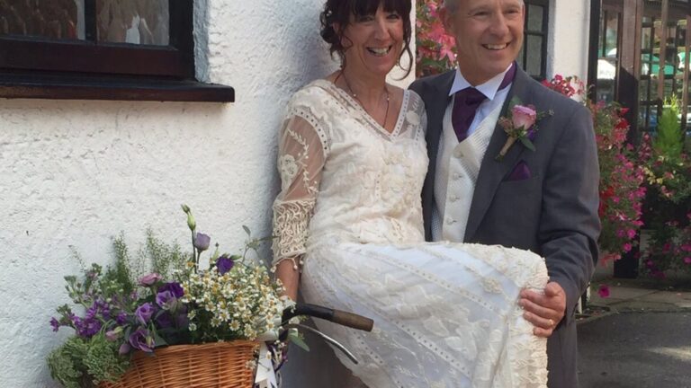 Couple outside a cottage, the bride in an ivory lace dress, Lesley Cutler Bridal Wear