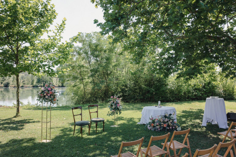 Lakeside ceremony set-up with chairs and a floral stand under the trees, Letizia T. Events