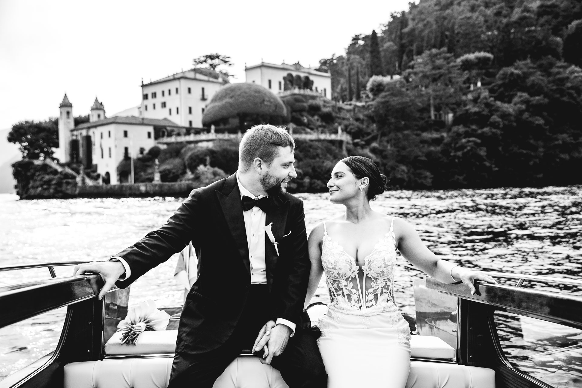 In black and white: the couple on a wooden launch, Villa del Balbianello on its headland behind, Lake Como, Light & Dreams