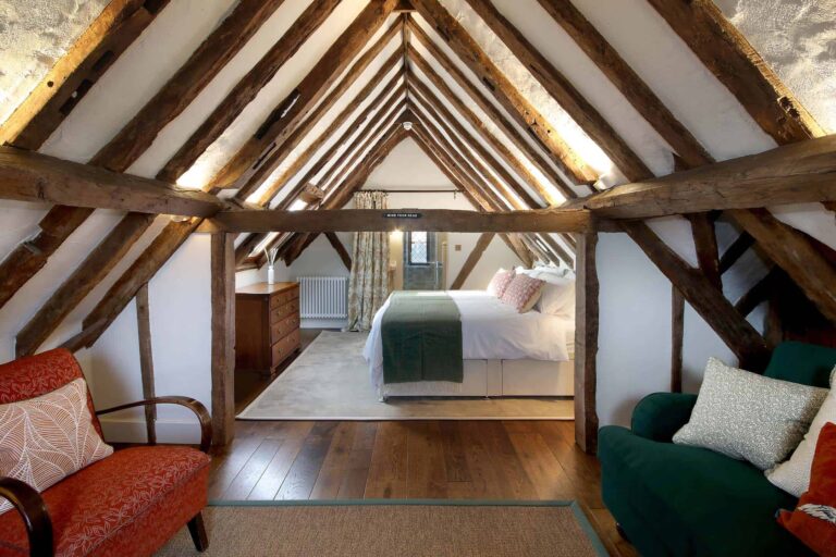 An attic bedroom under exposed oak trusses, with armchairs at one end and a double bed at the other, Lillibrooke Manor
