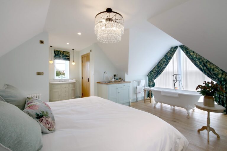 The Loft honeymoon suite: a bed under a sloping ceiling and a freestanding roll-top bath by the window, Lillibrooke Manor