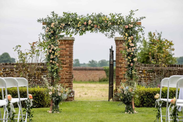 A floral arch between brick piers at the walled garden opening, with white chairs set either side of the aisle, Lillibrooke Manor