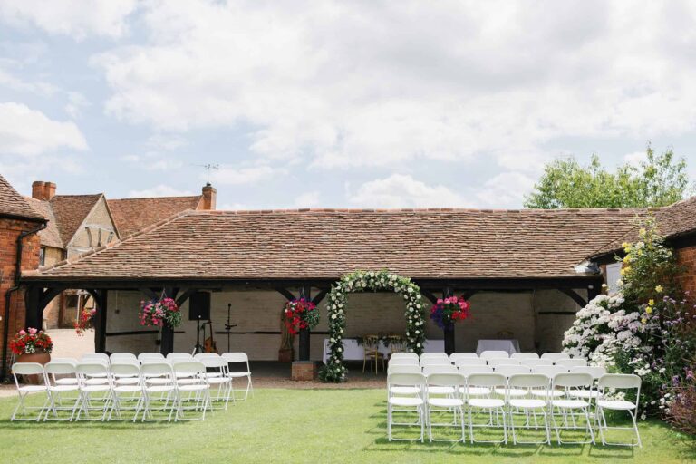 The covered cloister set for an outdoor ceremony, with white chairs on the lawn and a floral arch at the front, Lillibrooke Manor