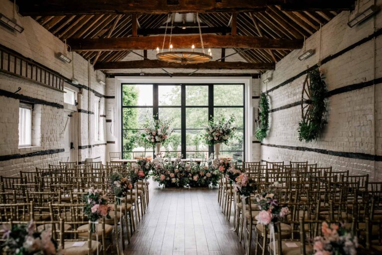 A whitewashed ceremony barn laid out with rows of chairs, a central aisle and a glazed end wall dressed with flowers, Lillibrooke Manor