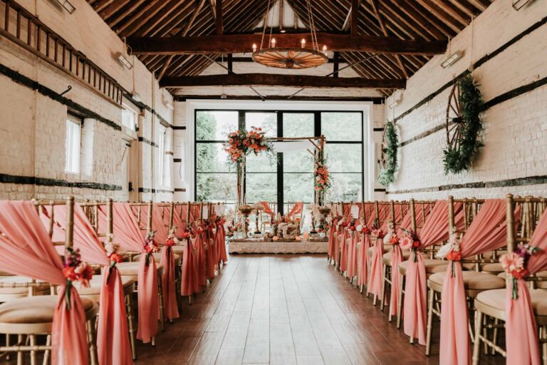 The same whitewashed ceremony barn dressed with pink chair sashes and dried flowers, under a wooden chandelier, Lillibrooke Manor