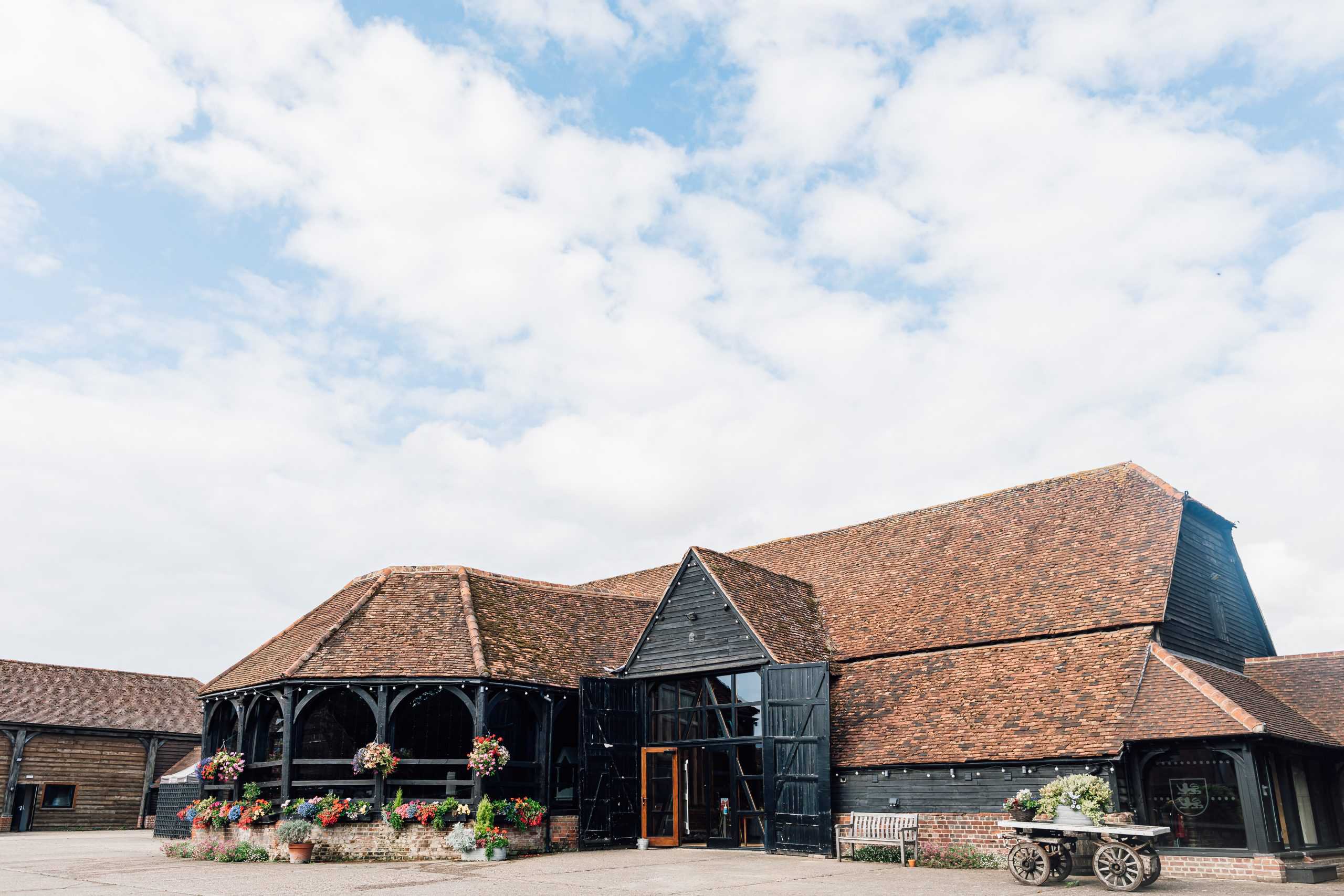The Great Barn from the forecourt: a long tiled roof, black weatherboarding and the glazed hexagonal Engine House on the left, with hanging baskets along the wall, Lillibrooke Manor