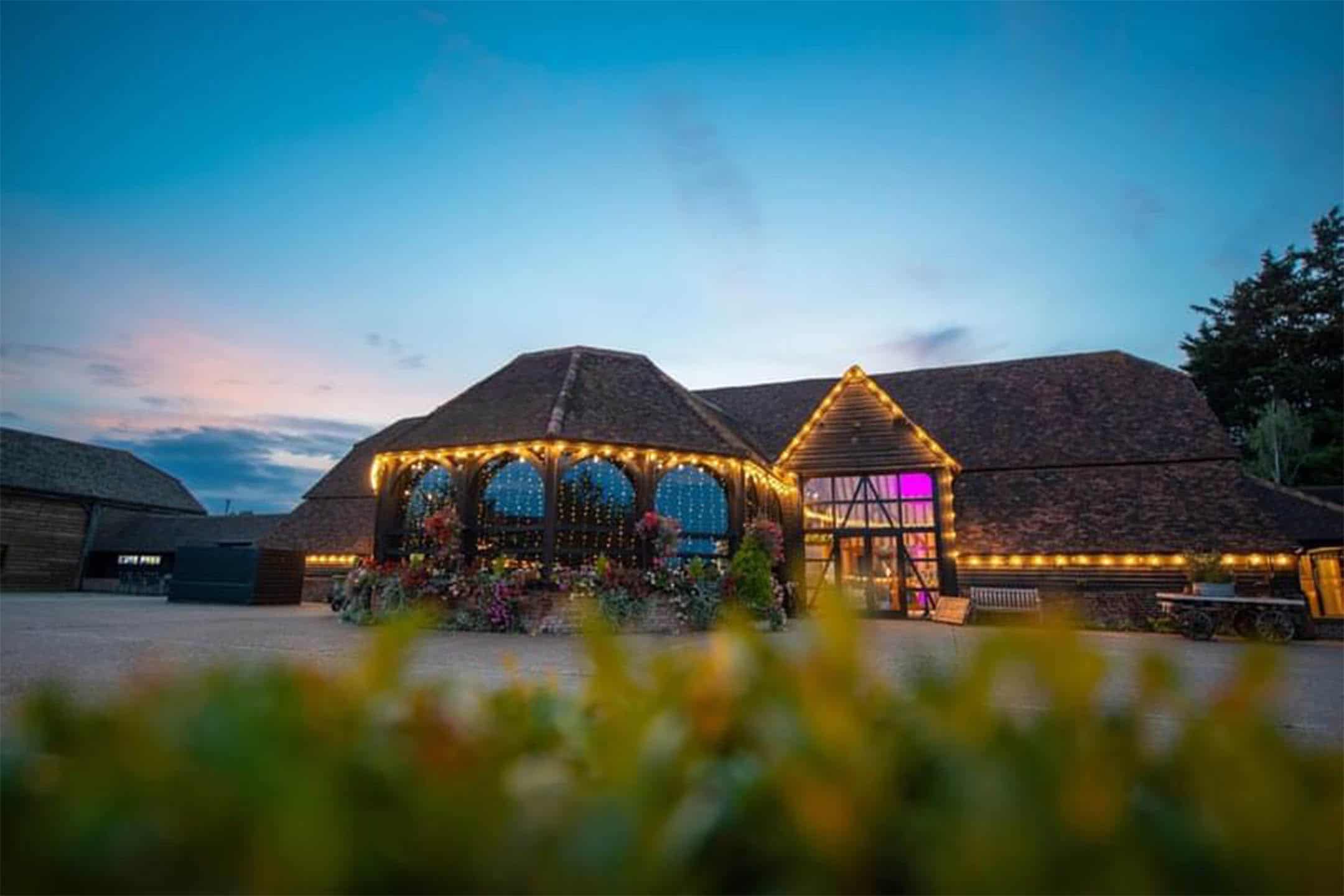 The Great Barn and Engine House at dusk, edged with festoon lights under a pink and blue sky, Lillibrooke Manor