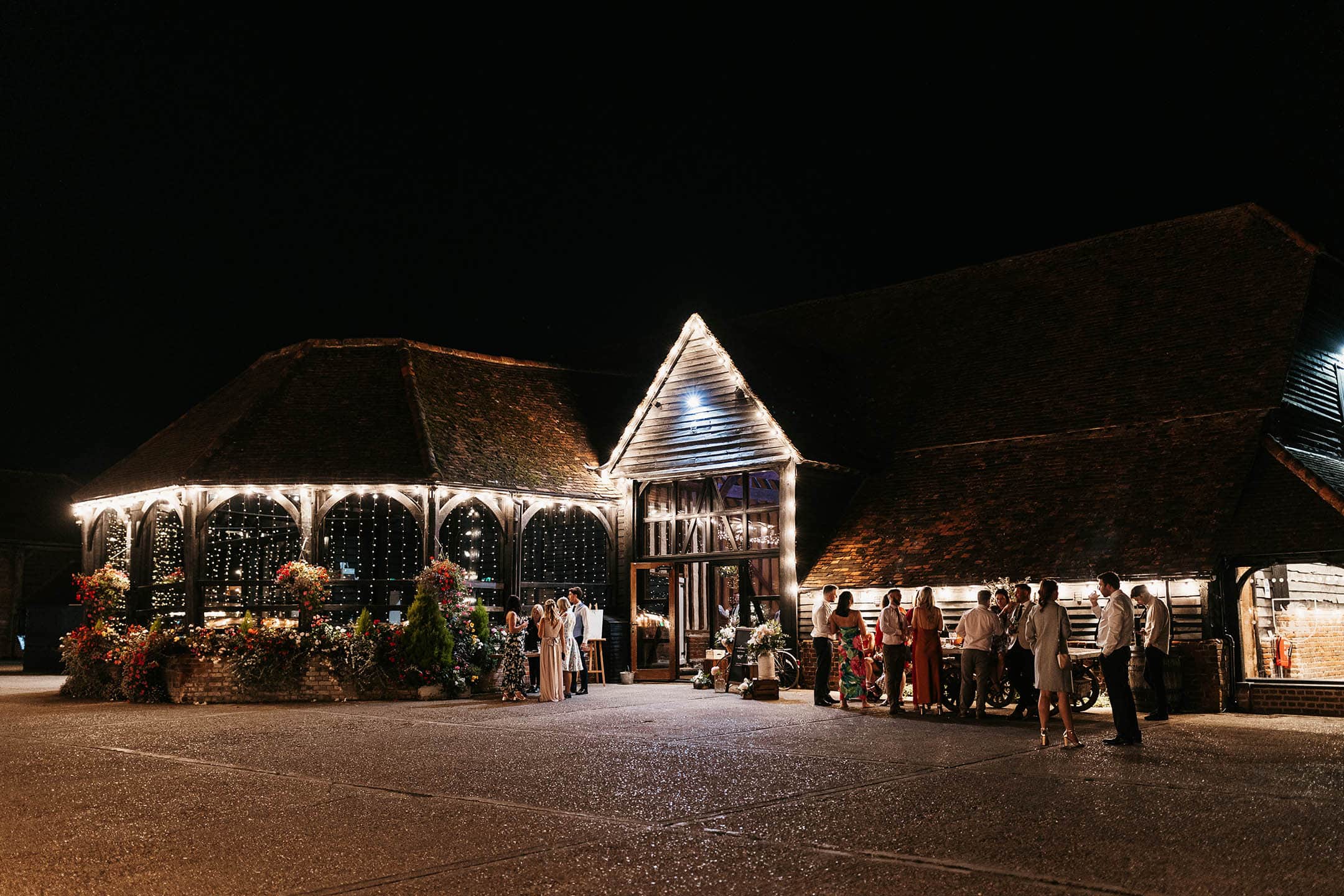 The forecourt at night, with the lit Engine House and the gabled entrance of the Great Barn, Lillibrooke Manor