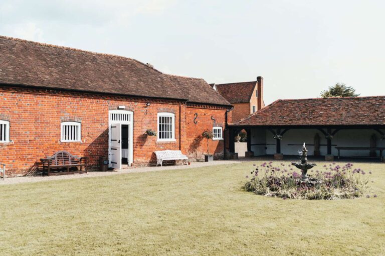Red-brick estate buildings around a lawn with a stone fountain and a planted bed, Lillibrooke Manor