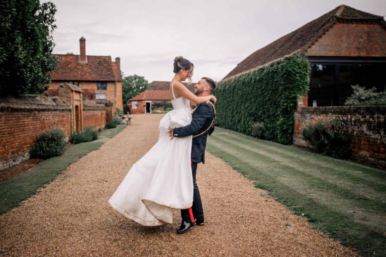 A couple on the gravel path beside clipped hedges, with the Manor House chimneys and a barn roof behind, Lillibrooke Manor