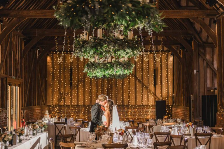The Great Barn set for the wedding breakfast, with a greenery chandelier, fairy-light curtain and round tables, Lillibrooke Manor