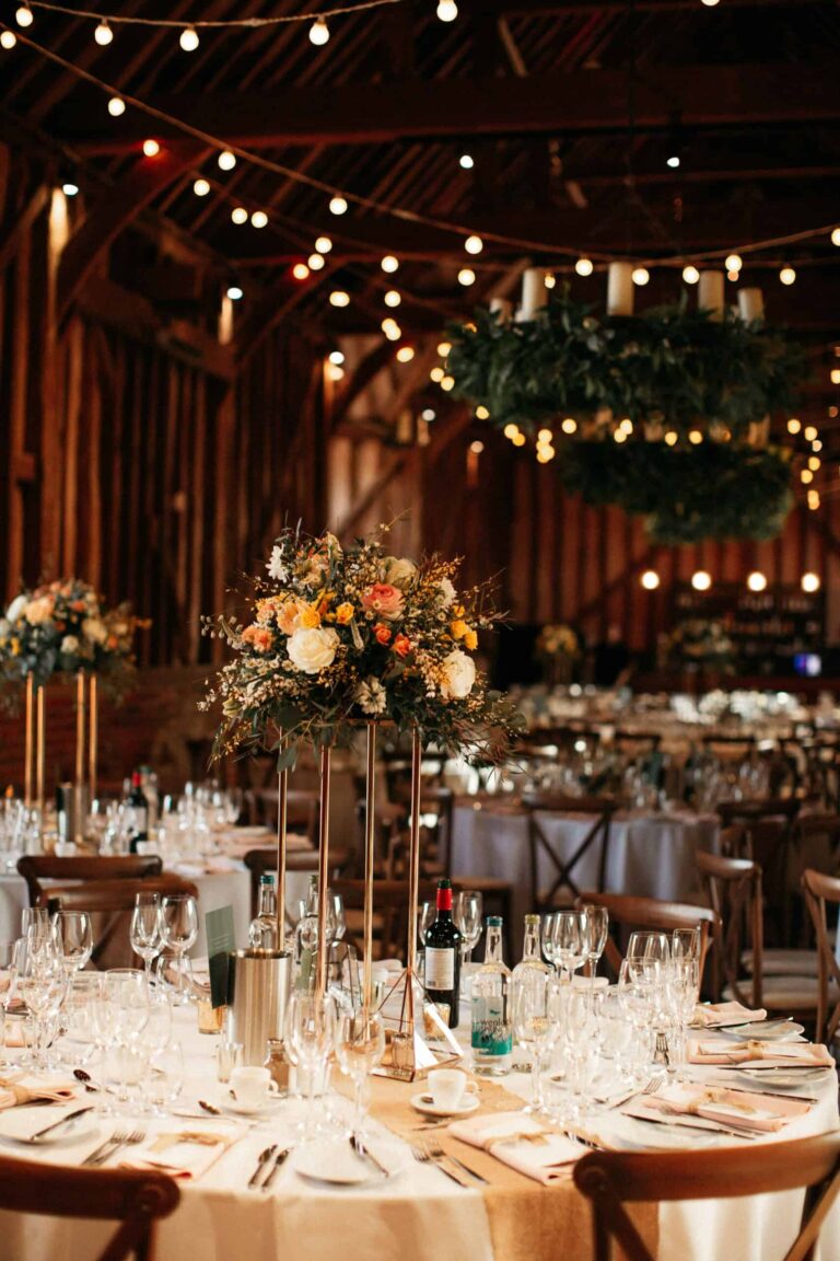 Round tables in the Great Barn under festoon lights and timber trusses, with tall floral centrepieces, Lillibrooke Manor