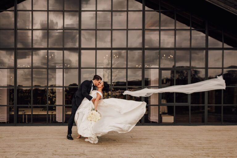 A dip and a kiss as the veil catches the wind outside a glass-walled venue, Lina + Tom Photography