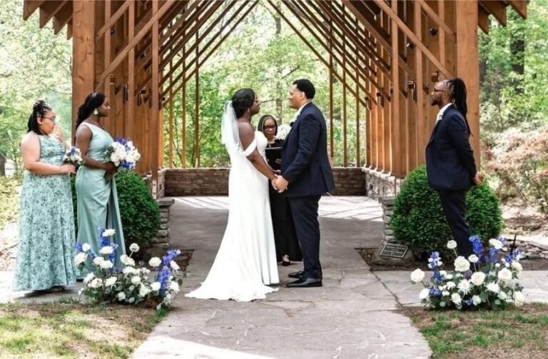 A ceremony under a timber-framed canopy with blue and white floral arrangements and guests in the trees beyond, Living Ceremonies