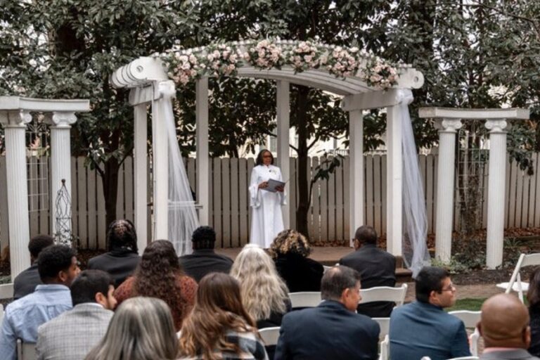 A garden ceremony beneath a white columned arbor decked with flowers, guests seated in rows, Living Ceremonies