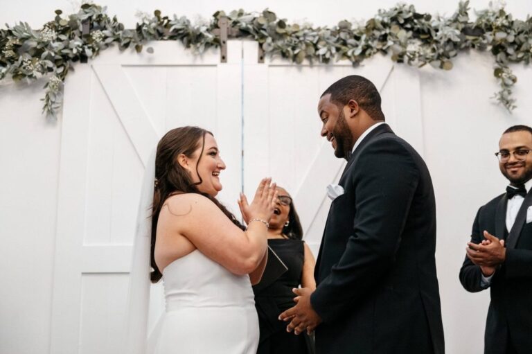 The couple laughing during their vows in front of white barn doors topped with greenery, Living Ceremonies