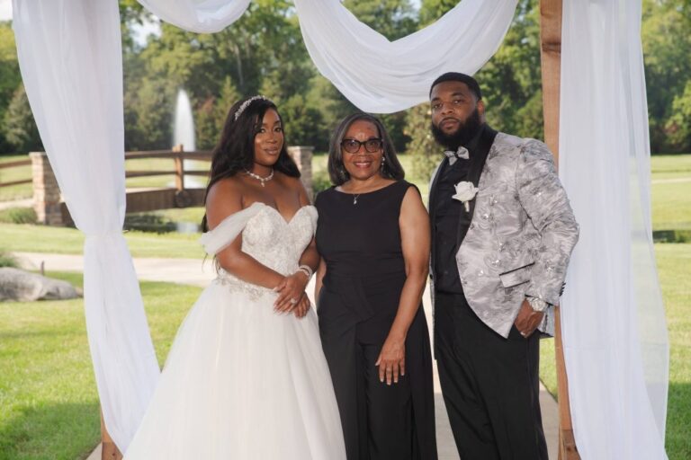 A couple and their officiant under white draped fabric on a lawn with a fountain behind, Living Ceremonies