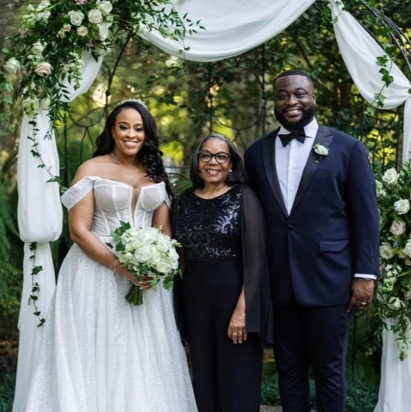 A bride and groom with their officiant beneath a floral arch of greenery and white blooms, Living Ceremonies
