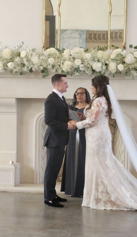 A couple exchanging vows at a stone fireplace dressed with white roses and candles, Living Ceremonies