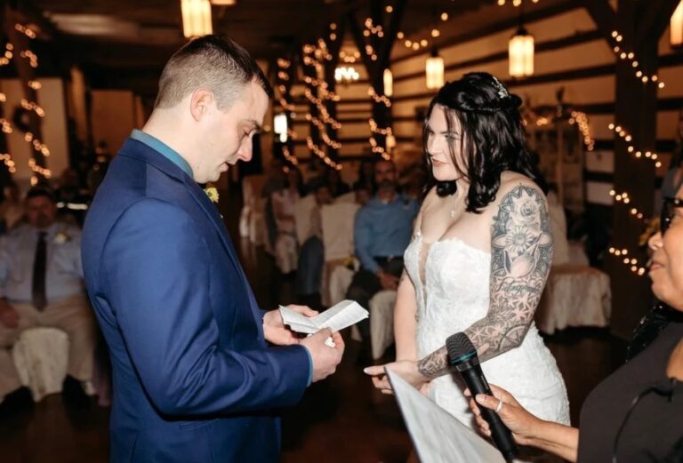A groom reading to his bride at the reception, with string lights and guests behind, Living Ceremonies