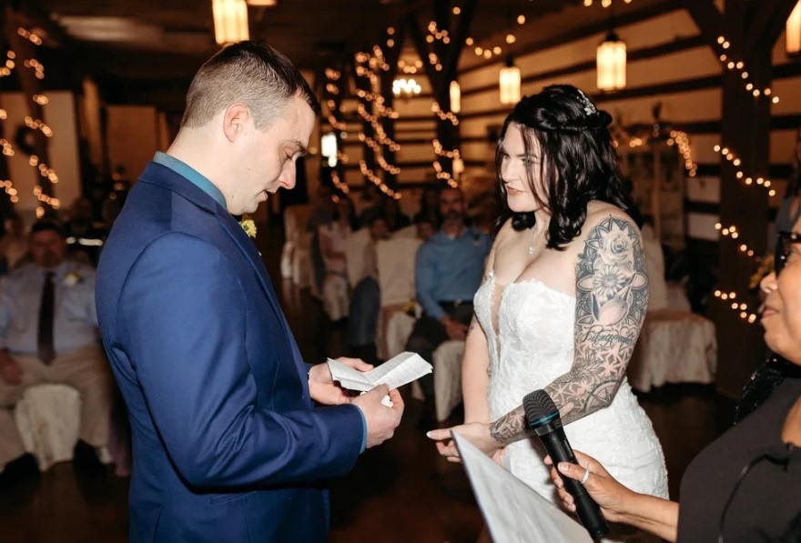 A groom reading to his bride at the reception, with string lights and guests behind, Living Ceremonies