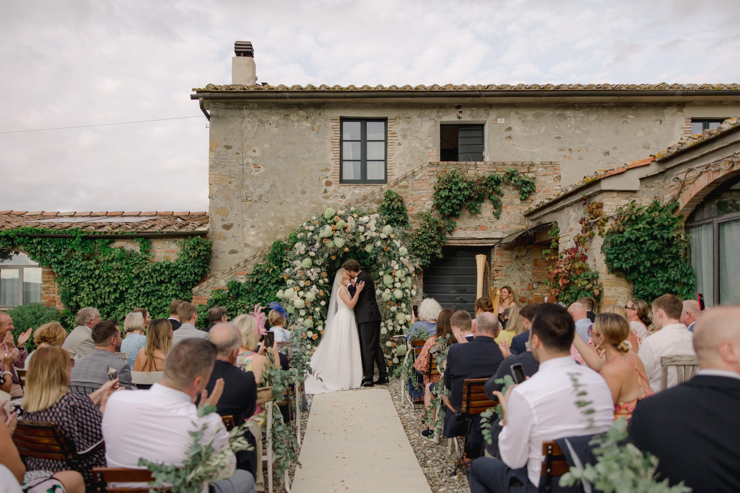 A ceremony in front of the farmhouse, the couple beneath a floral arch with guests seated either side, Locanda in Tuscany