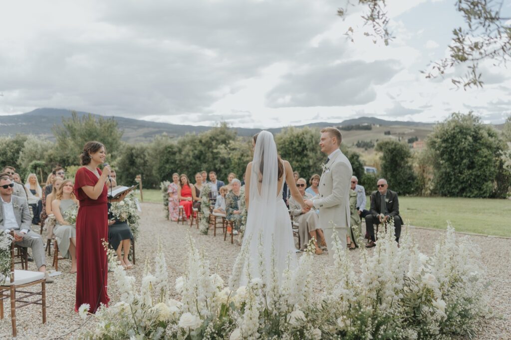 The couple and celebrant at an outdoor ceremony, guests seated in front of white flowers, Locanda in Tuscany