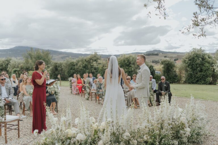 The couple and celebrant at an outdoor ceremony, guests seated in front of white flowers, Locanda in Tuscany