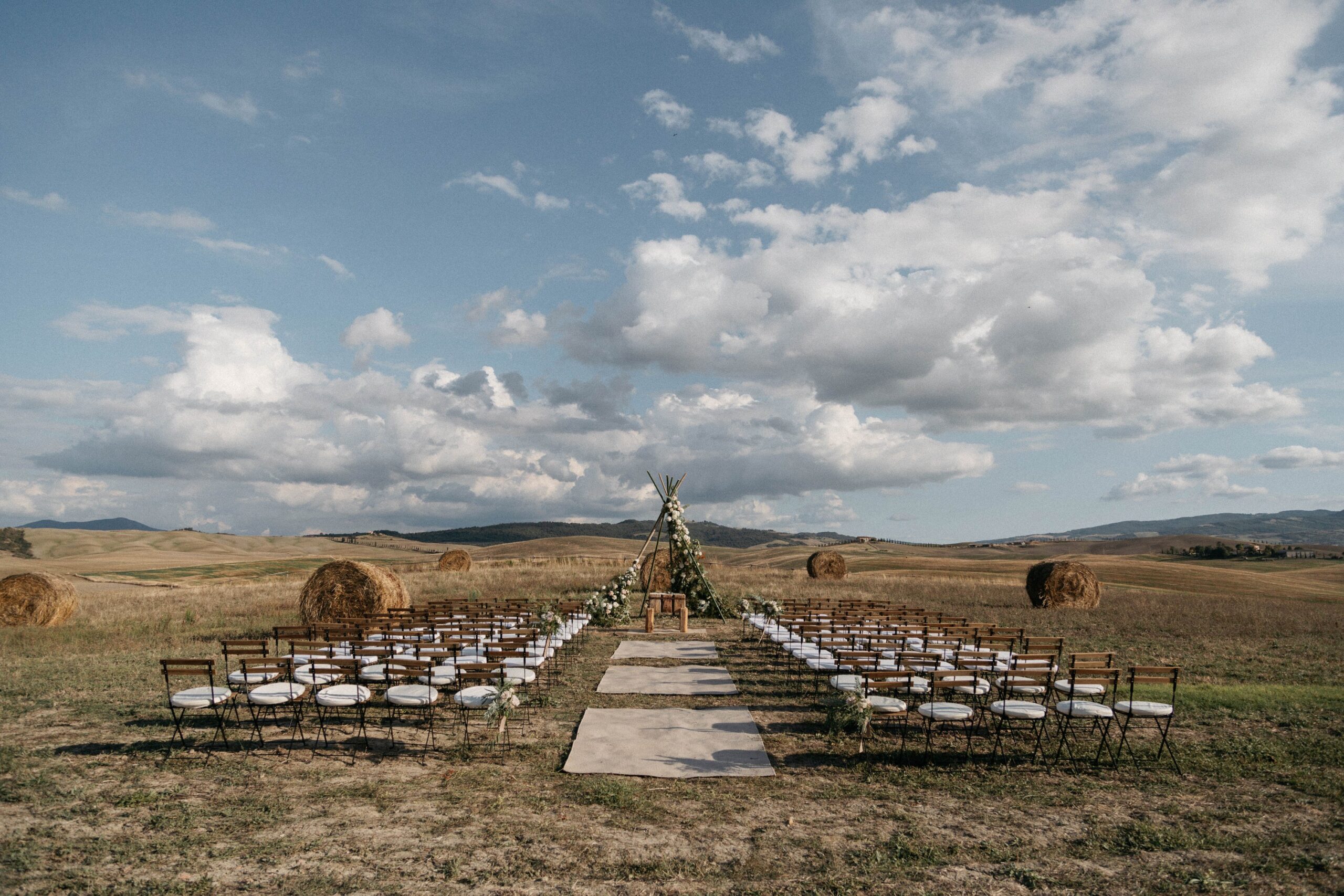 A ceremony aisle between rows of chairs in the open fields, with hay bales beyond