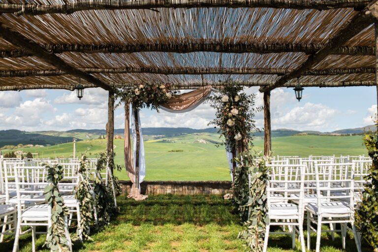 A floral arch and white chairs beneath the reed-roofed pergola, looking out over green fields, Locanda in Tuscany