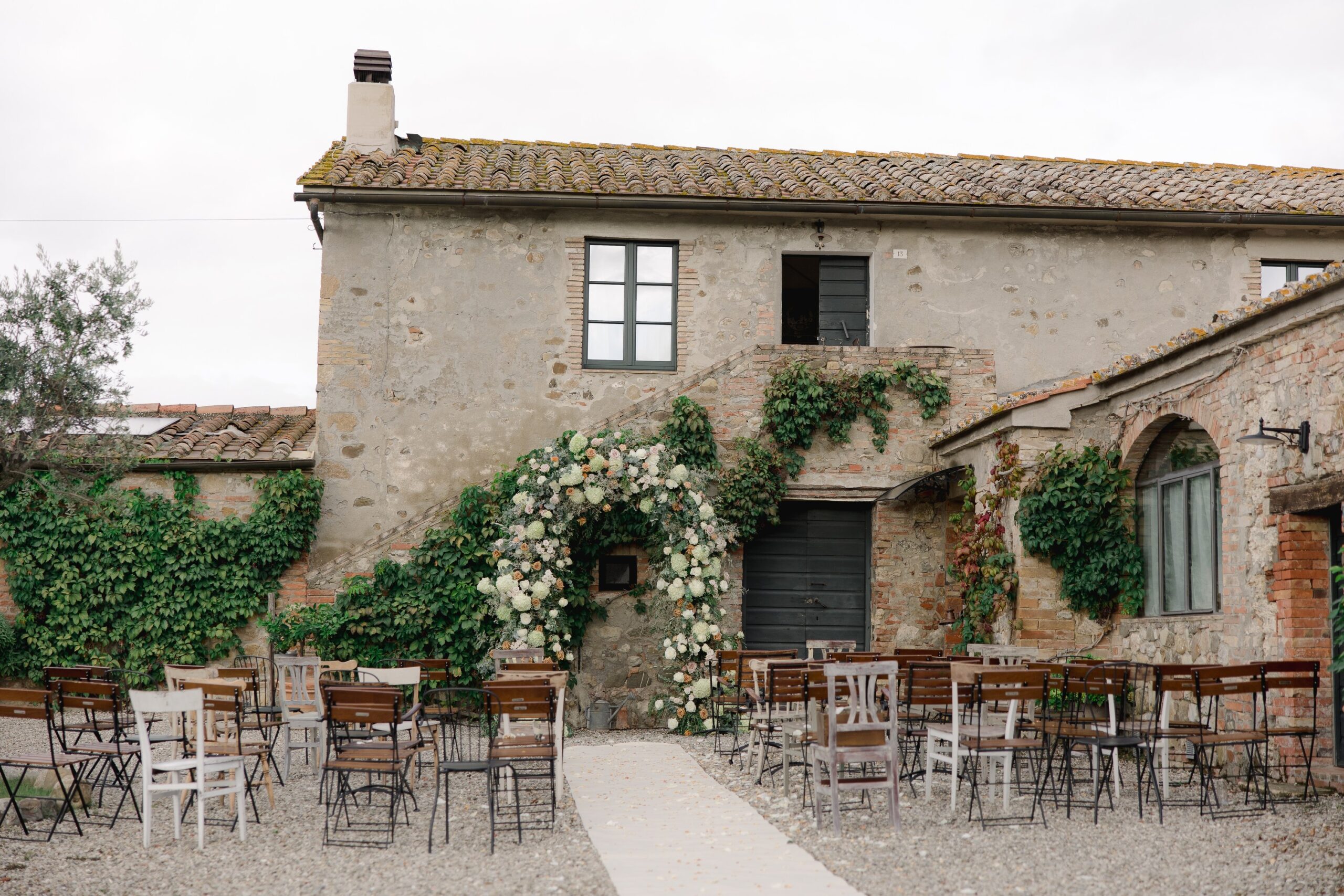 The stone farmhouse with a floral arch and ceremony chairs set out on the gravel courtyard, Locanda in Tuscany