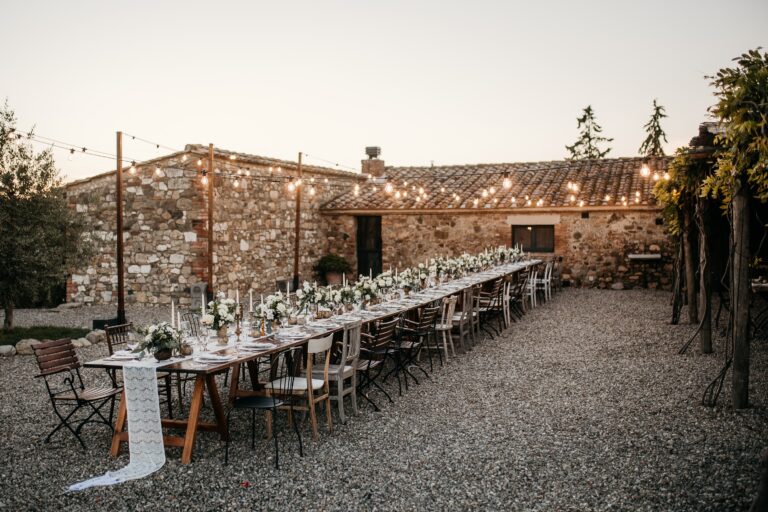 A long table in the gravel courtyard beside the stone building, strung with festoon lights, Locanda in Tuscany
