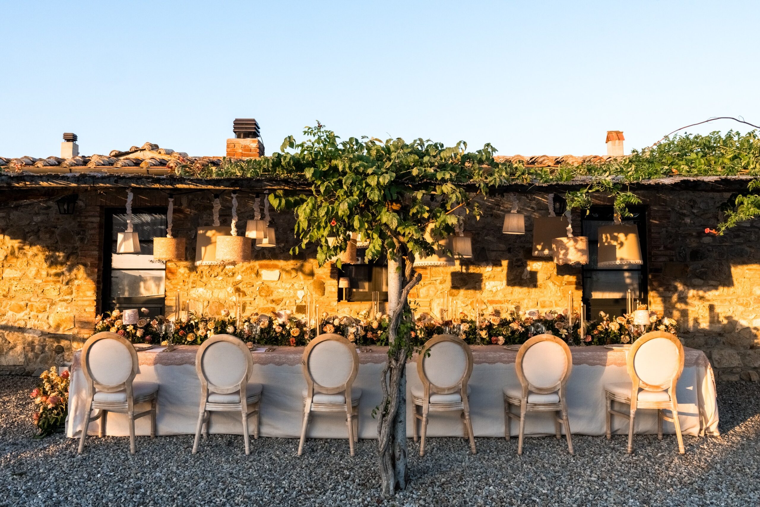 A long dinner table with a vine overhead in front of the stone building at golden hour