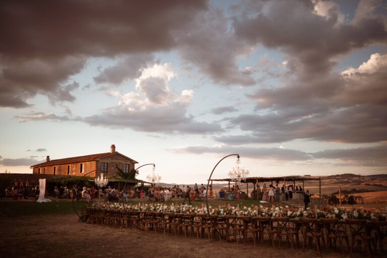 A long table laid in the open field at dusk, the farmhouse behind, Locanda in Tuscany