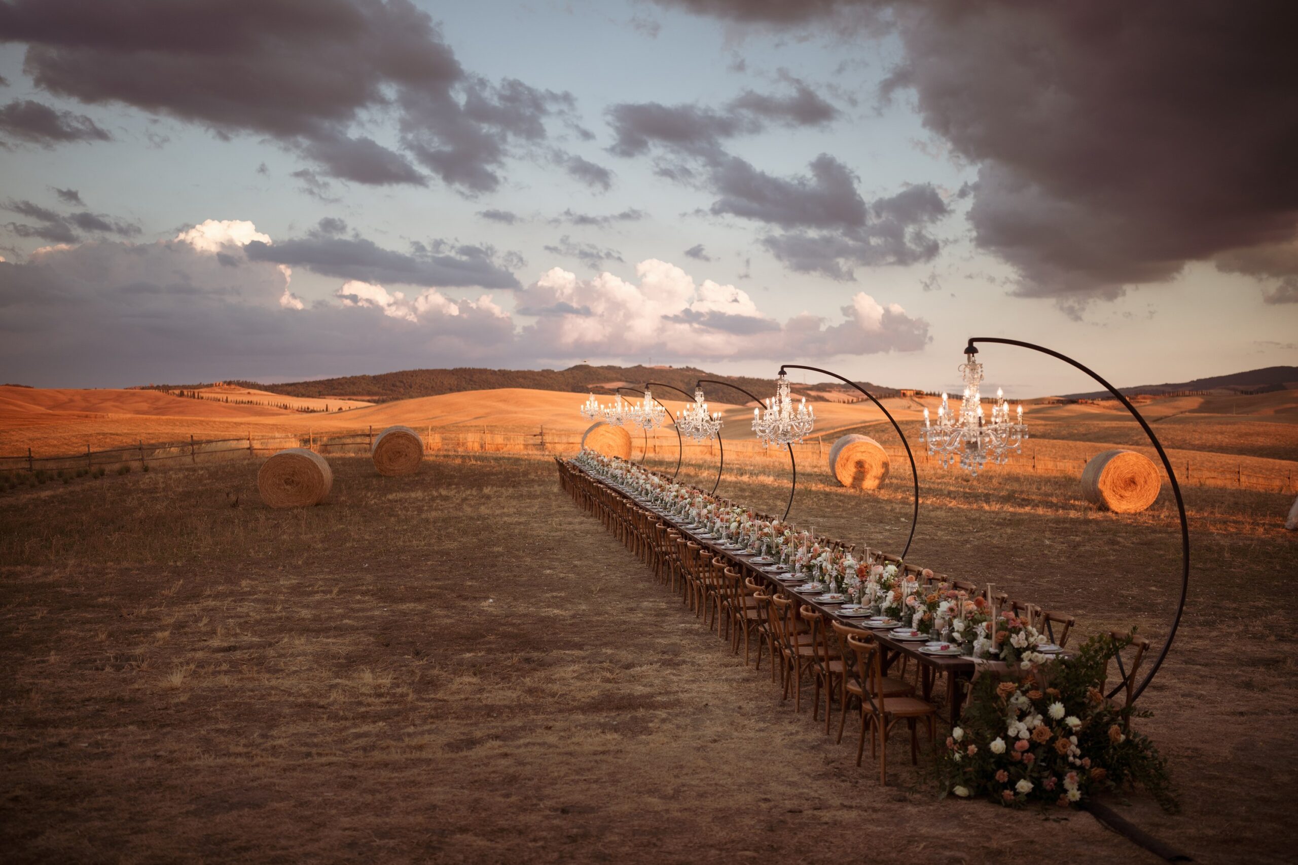 A long dinner table with chandeliers on curved stands in a field at sunset