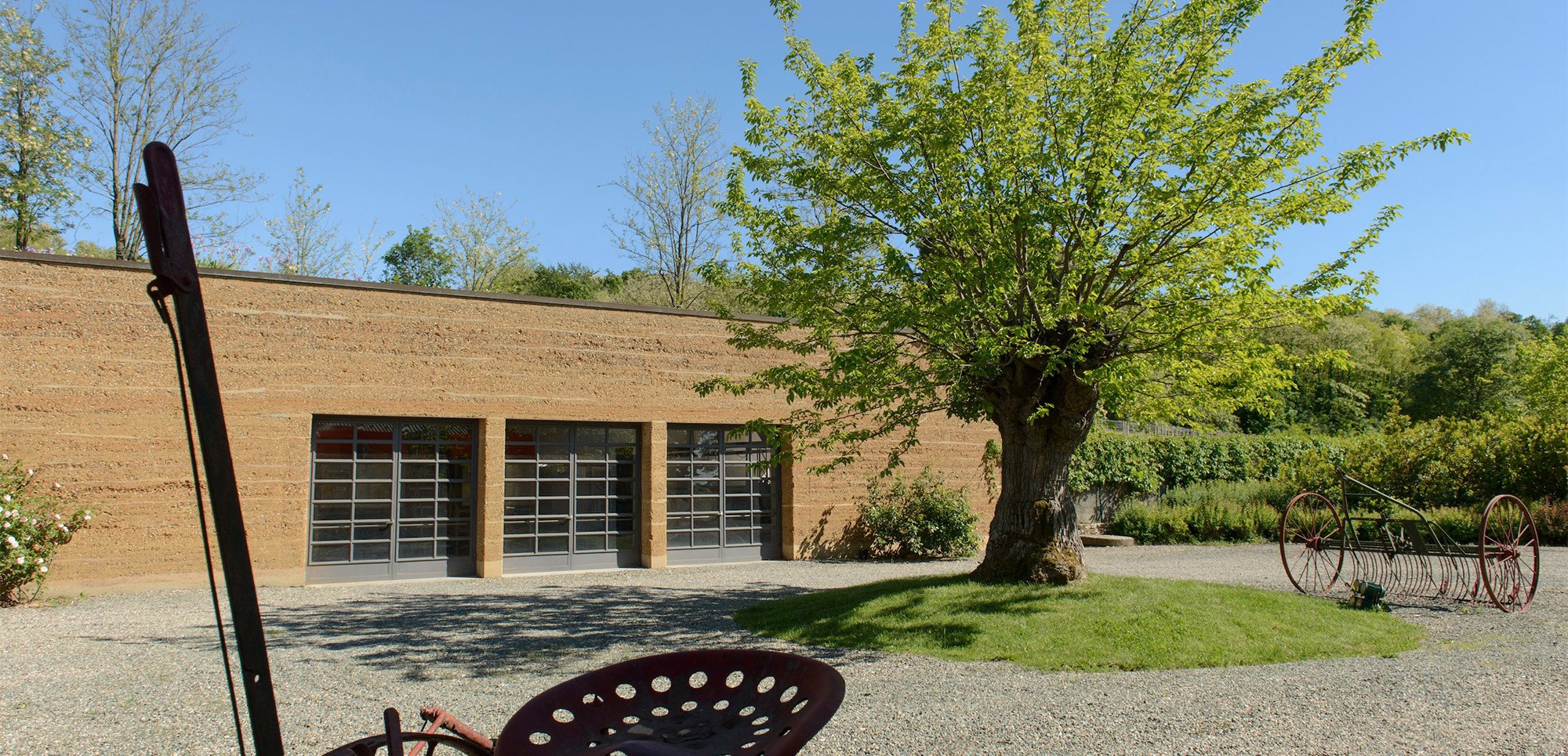 A rammed earth building with glass doors and a tree in a gravel yard on the La Raia estate