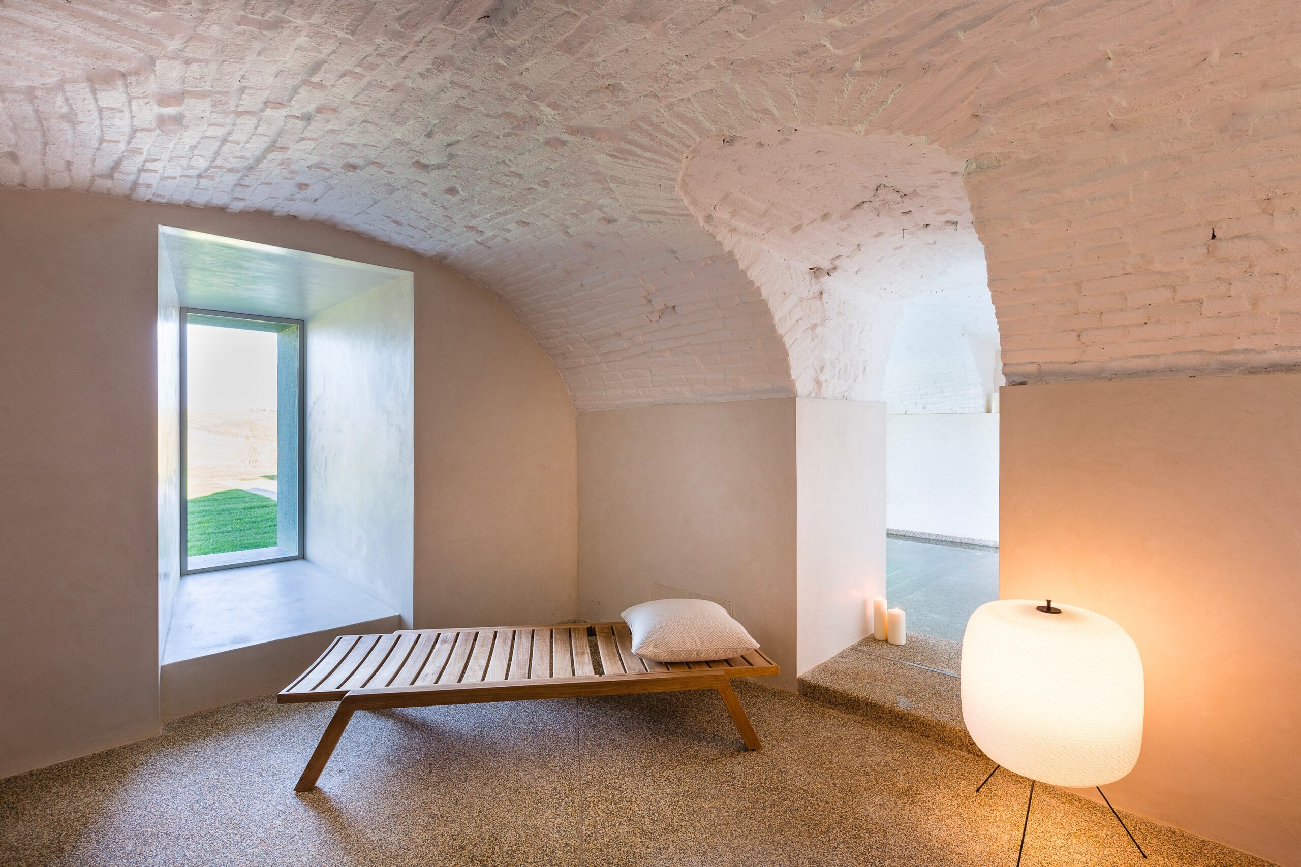 A vaulted white room with a wooden daybed and a lantern lamp beside a window