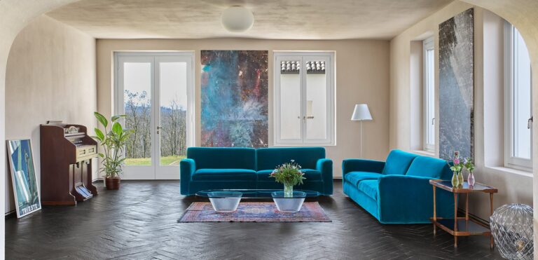 A sitting room with a teal sofa, a piano and large paintings, Locanda La Raia