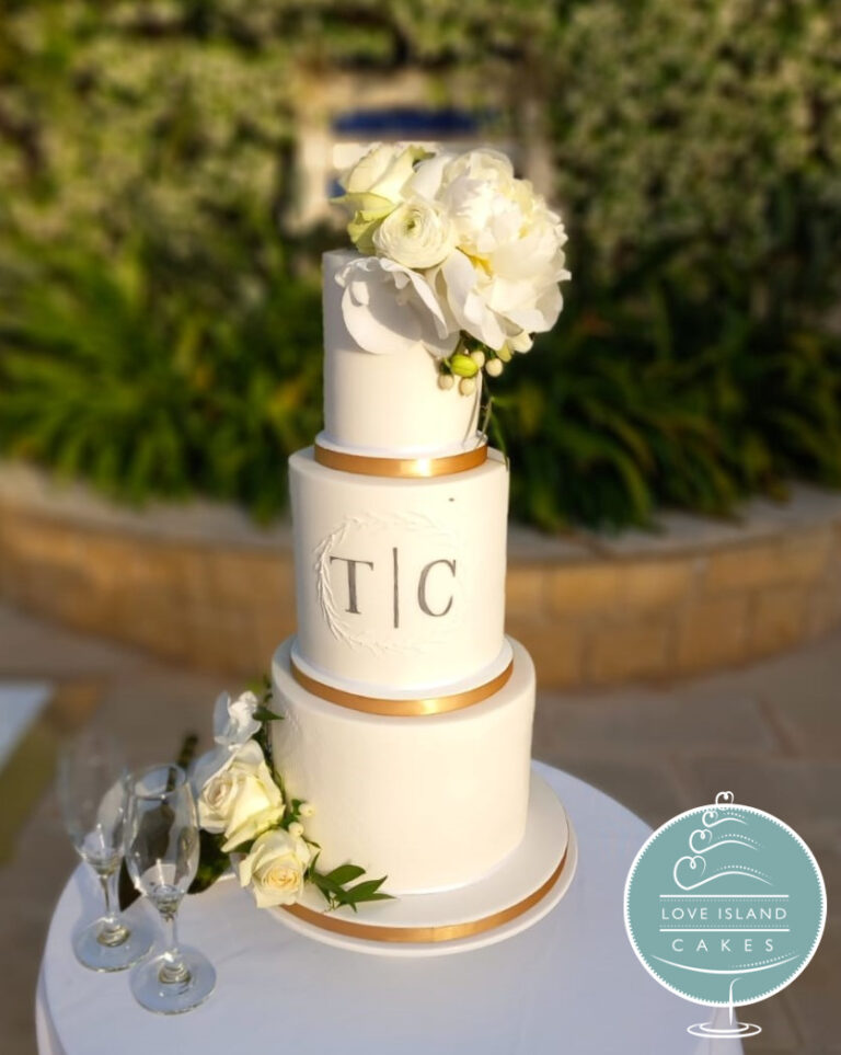 A white tiered cake with a TC monogram and gold bands, with a white flower crown, set up outdoors with champagne flutes, Love Island Cakes