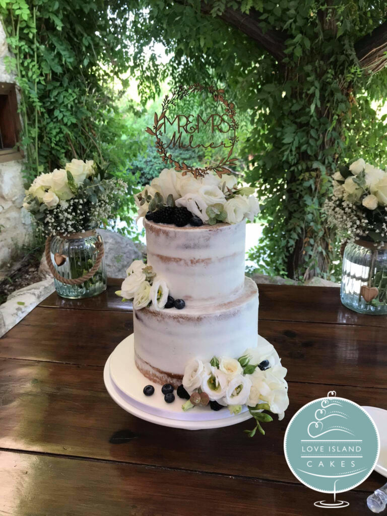A semi-naked two-tier cake with white flowers and berries on a wooden table beneath a vine pergola, Love Island Cakes