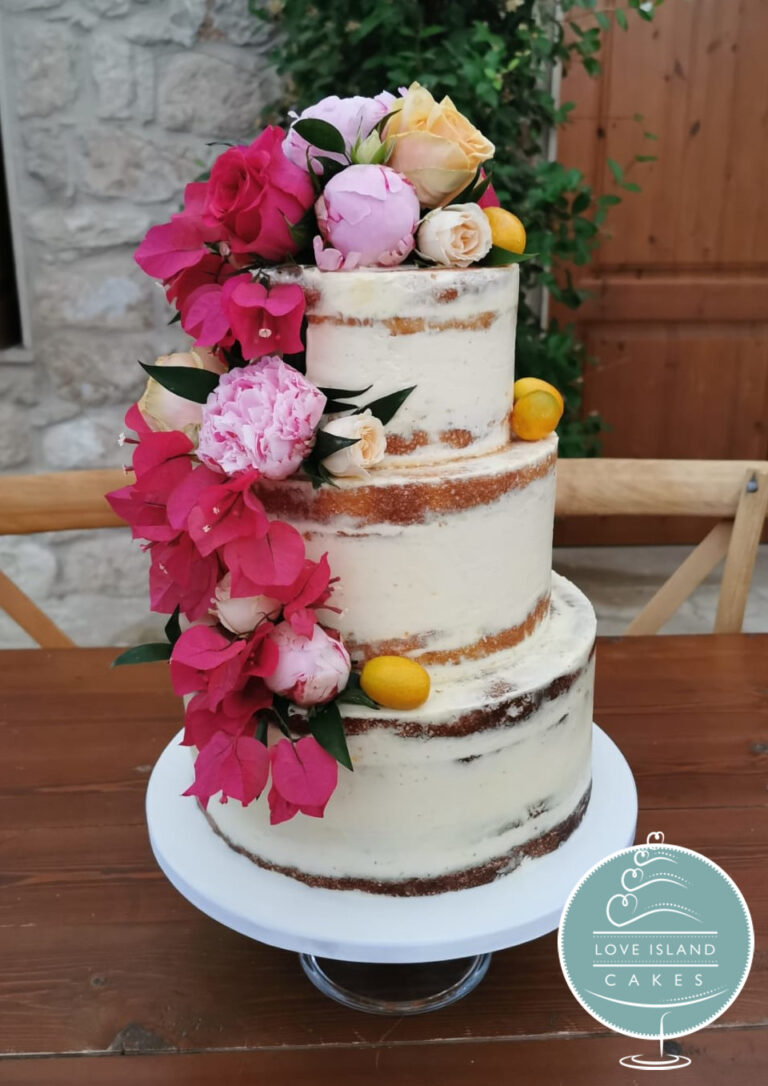 A three-tier semi-naked cake dressed with bright pink blooms and lemons on a white stand, stone wall behind, Love Island Cakes