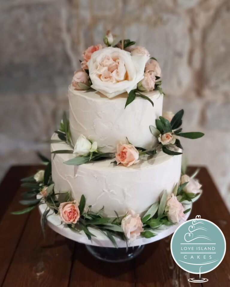 A white textured two-tier cake with blush roses and olive leaves on a wooden table, Love Island Cakes