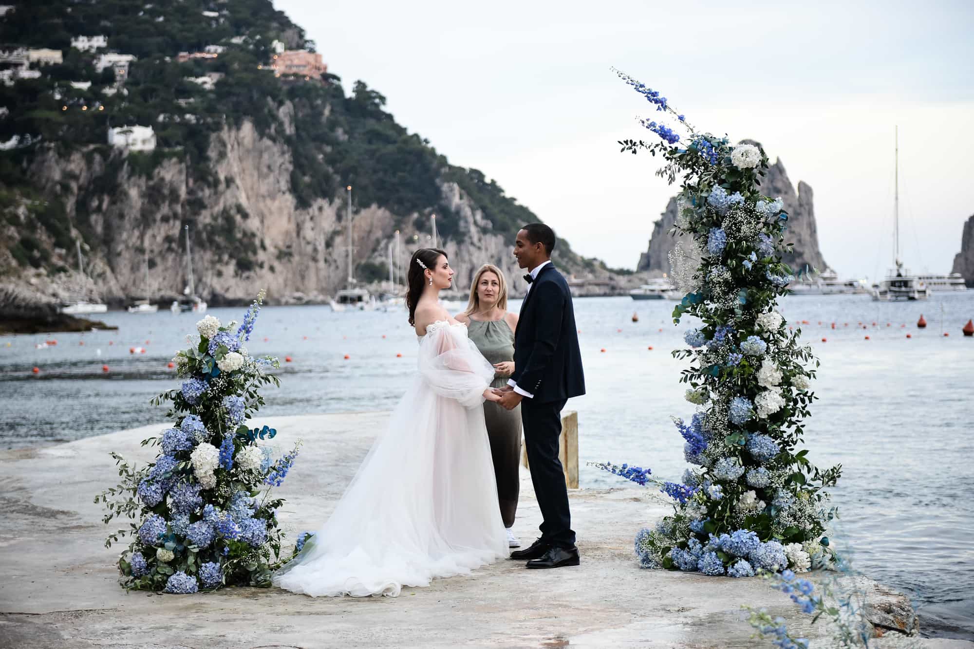 A ceremony on the rocks by the sea on Capri, between two blue hydrangea columns with the Faraglioni behind; a styled wedding shoot organised by Say Yes In Italy, Lovexposed Photography