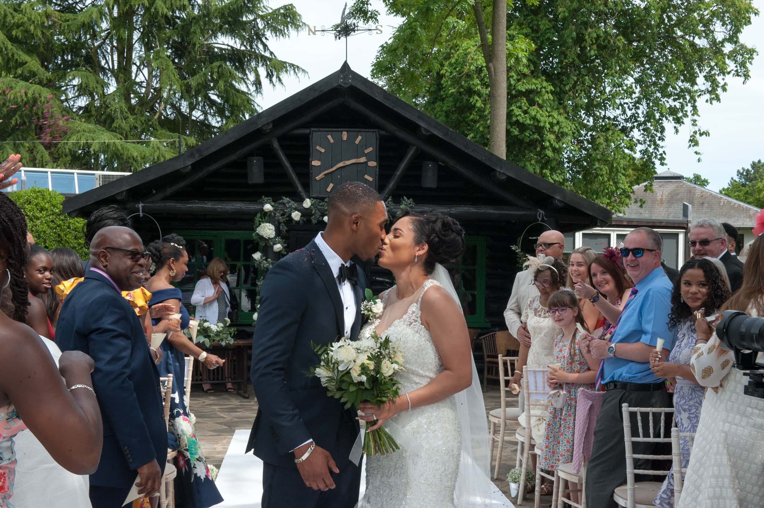 The first kiss at the end of the aisle at an outdoor ceremony, guests either side; Joelle and Tashan's wedding at Gilwell, Essex, Lovexposed Photography