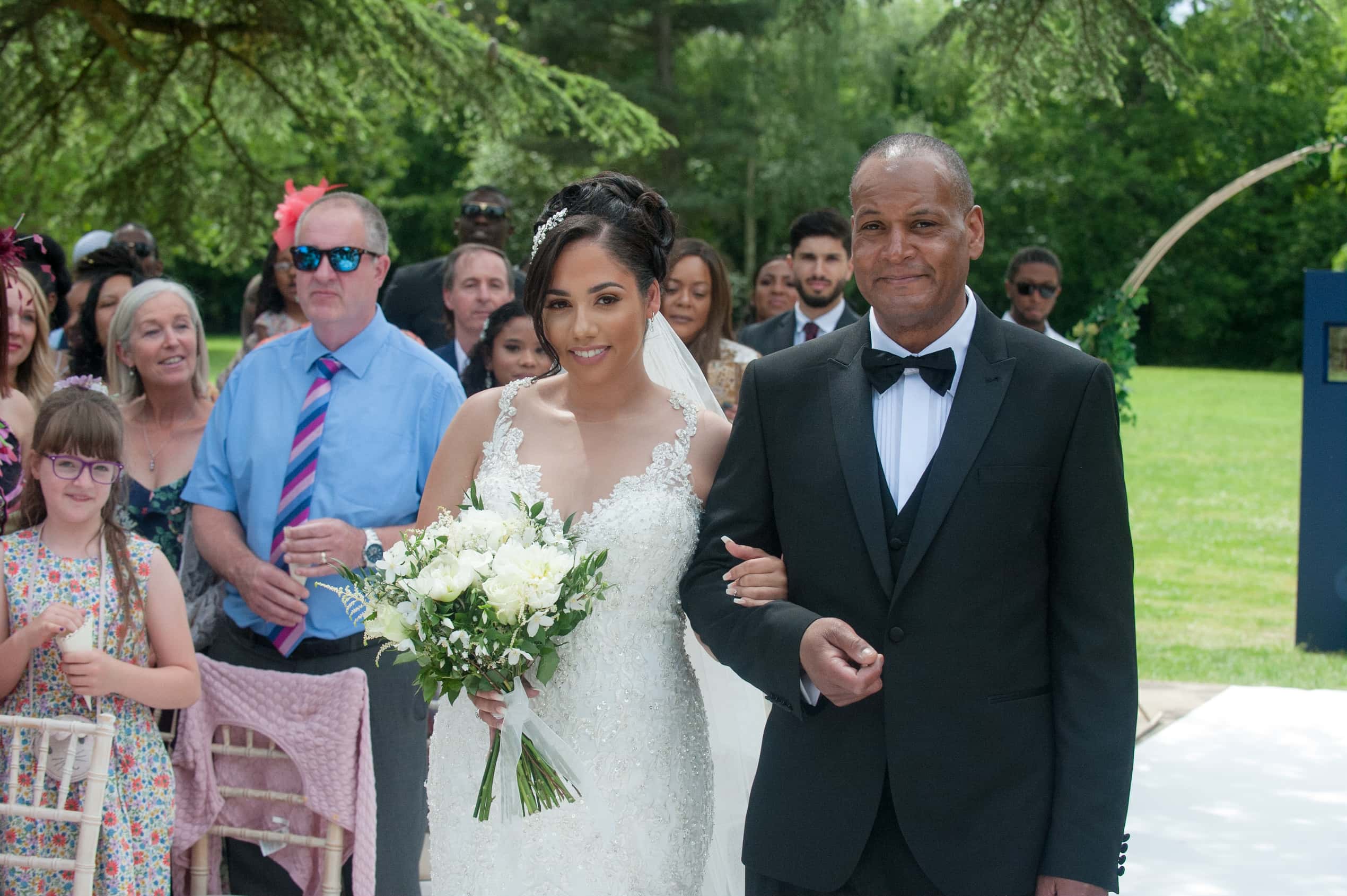 The bride walks down the outdoor aisle with her father; Joelle and Tashan's wedding at Gilwell, Essex, Lovexposed Photography